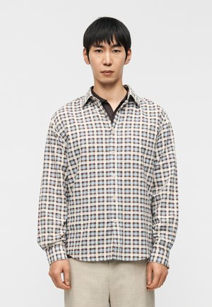 Uomo che indossa una camicia a quadri marrone, blu e bianca sopra una polo scura, abbinata a pantaloni beige chiaro, in piedi davanti a uno sfondo semplice.