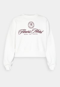 Felpa bianca corta con scollatura rotonda, caratterizzata da un logo testuale bordeaux e un'illustrazione di un fiore, che enfatizza i servizi di hotel e spa.