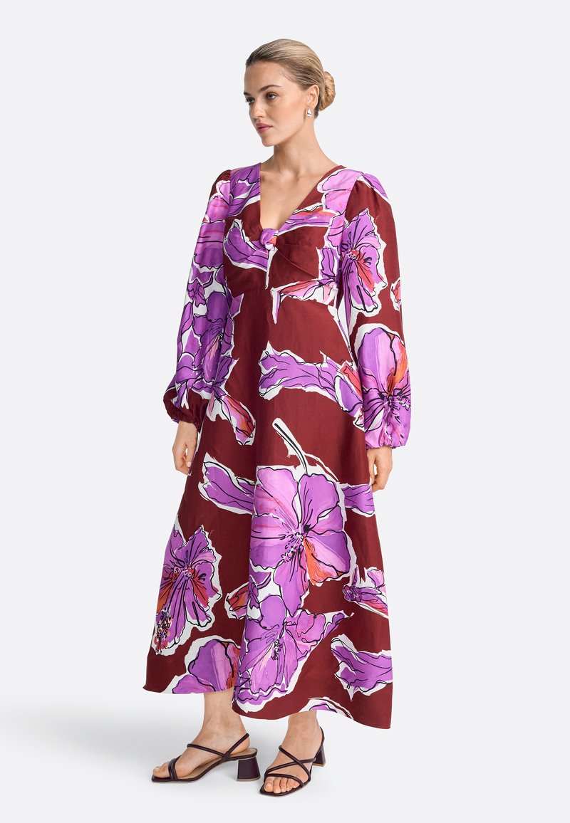 Naine blondide juustega, kootud kuklas, kannab pikkade varrukatega, pahkluuni ulatuvat burgundi punast kleiti suurte roosade ja lillade lillemustritega ning musti kontsakingi.