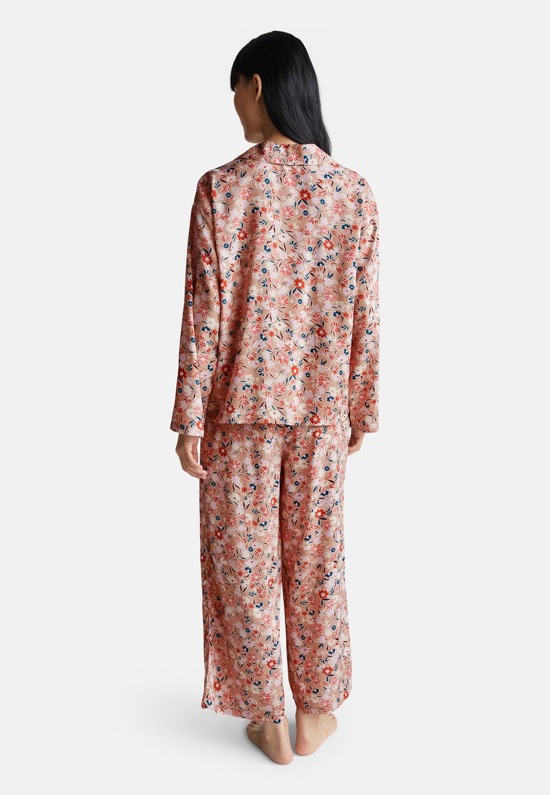 Donna in piedi a piedi nudi, di spalle, che indossa un comodo pigiama floreale rosa con maniche lunghe e pantaloni larghi, su uno sfondo semplice.