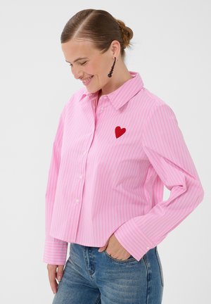 Femme souriante portant une chemise rose à rayures boutonnée avec un patch en forme de cœur rouge sur la poitrine et un jean bleu, main dans la poche.