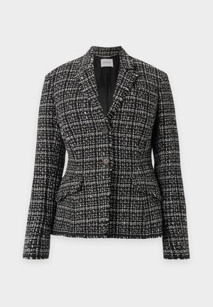 Kvinde stående iført en sort-hvid ternet blazer, sort top, brede hvide bukser og sorte sko mod en ensfarvet baggrund.