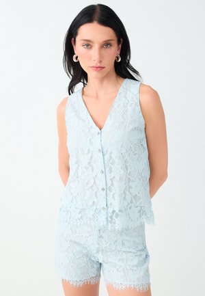 Donna con capelli scuri indossa un top in pizzo azzurro chiaro senza maniche e pantaloncini abbinati, accessoriata con grandi orecchini a cerchio, in piedi su sfondo bianco.