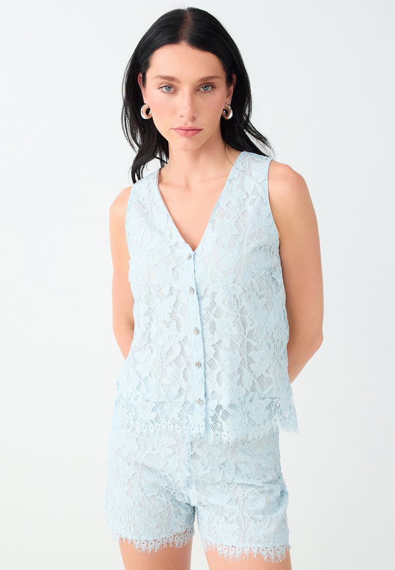 Femme aux cheveux foncés portant un haut en dentelle bleu clair sans manches et un short assorti, accessoirisée de grandes boucles d'oreilles créoles, debout sur fond blanc.