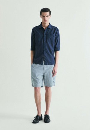 Joven de pie con las manos en los bolsillos, vistiendo una camisa azul marino de manga larga, pantalones cortos blancos y azules a rayas, y zapatos negros, sobre un fondo liso.