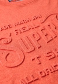 Coral cotton t-shirt with raised text design reading "SUPER" and "REAL" in bold, featuring a round neckline and soft texture.
