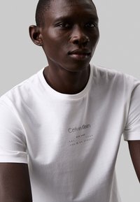 T-shirt bianco in cotone con scollatura a girocollo, caratterizzato da un sottile testo di branding grigio sul petto. Tessuto liscio, design aderente.
