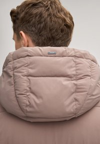 Giubbotto imbottito di un delicato colore rosa, con una texture morbida, caratterizzato da un alto colletto e un'etichetta metallica sul retro. Design liscio e minimale.