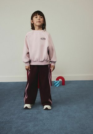 Barn, der står på blåt tæppe iført en lyserød sweatshirt og mørke bukser med pink striber, med røde og blå legetøj i baggrunden.