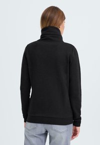 Černá mikina s vysokým límcem a dlouhými rukávy. Tkanina vypadá měkce a texturovaně, je přiléhavá v pase a má jednoduchý, minimalistický design.