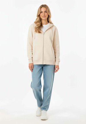 Felpa beige con zip e due tasche frontali, indossata sopra una maglietta bianca, abbinata a pantaloni in denim azzurro chiaro e sneakers bianche.