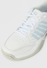 Sapatilha atlética branca com acentos em azul claro, apresentando um cabedal de couro liso, caixa de biqueira perfurada, atacadores planos e sola de borracha texturizada.