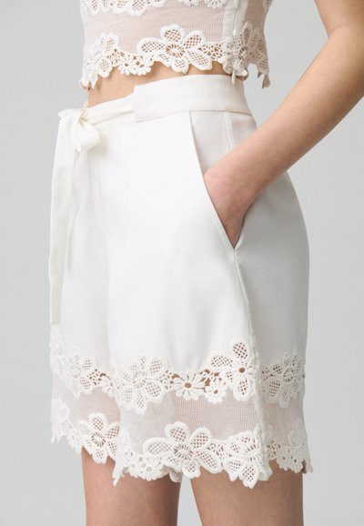 Pantaloni corti bianchi con pizzo decorativo, caratterizzati da motivi floreali, una texture liscia e una vita con lacci. Comprendono pratiche tasche laterali.