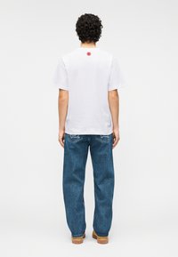 Λευκό βαμβακερό t-shirt με κόκκι νο κυκλικό λογότυπο στην πλάτη, συνδυασμένο με φαρδιά μπλε τζιν παντελόνια και καφέ παπούτσια.