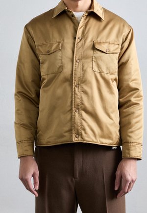 Homme portant une veste beige à boutons avec poches poitrine à rabat par-dessus une chemise blanche et un pantalon marron, debout devant un fond uni.