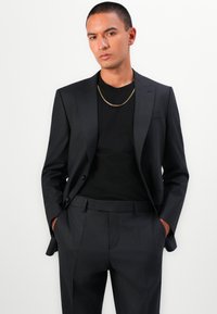 Schwarze Jacke mit strukturiertem Finish, kombiniert mit einem einfarbigen schwarzen T-Shirt und dunklen Hosen. Akzentuiert durch eine goldene Kette.