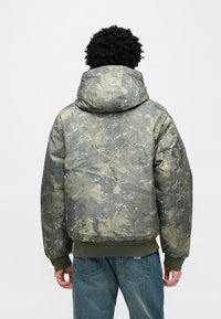 Maskáčová prešívaná bunda v zelených a béžových tónoch s kapucňou, rebrovanými manžetami a textúrovaným povrchom s motívmi listov.