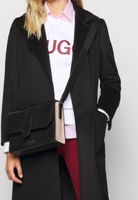 Svart handväska i krokodilpräglat läder med klaff, bruna detaljer och avtagbar axelrem. Bärs med en svart kappa över en vit sweatshirt.