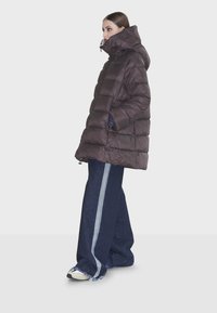 Giubbotto puffer marrone con colletto alto, caratterizzato da un design trapuntato e tasche con zip, abbinato a pantaloni in denim a gamba larga con strisce laterali.