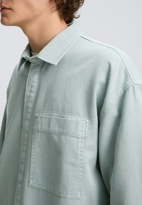 Lichtgroen, geribbeld shirt met een kraag, een enkele borstzak en zichtbare stiksels. De stof lijkt textuur te hebben en is iets oversized.