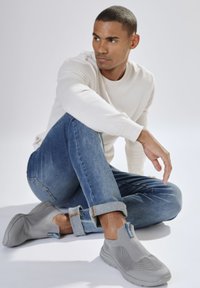 Junger Mann, der mit einem Bein angewinkelt auf dem Boden sitzt, trägt einen beigen Pullover, hellblaue hochgekrempelte Jeans und graue Slipper.