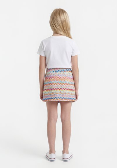 Παιδί στέκεται γυρισμένο με την πλάτη, φοράει λευκό μπλουζάκι με κοντά μανίκια, πολύχρωμη φούστα με ζιγκ-ζαγκ μοτίβο και λευκά αθλητικά παπούτσια σε απλό φόντο.