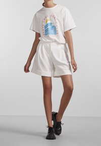 Bílý grafický bavlněný tričko s designem vlny a textem "Santa Cruz Beach Club" v kombinaci s vysokými bílými šortkami a černými teniskami.