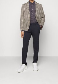 Beige geblokte blazer over een marineblauw gestippeld shirt, gecombineerd met donkere gepaste broek en witte sneakers. Opmerkelijke details zijn de knopen op de blazer.