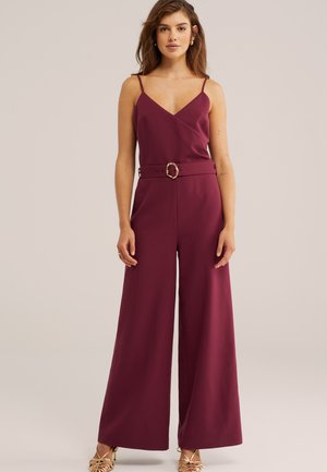 Femeie purtând un combinezon fără mâneci, de culoare burgund închis, cu picioare evazate și o curea asortată cu un cataramă aurie, stând în picioare pe un fundal simplu.