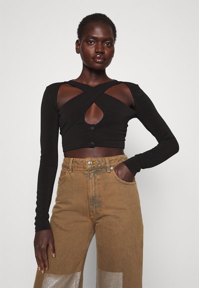 Top corto negro de manga larga con diseño entrecruzado y cierre de botón; combinado con jeans de mezclilla marrones de talle alto que presentan acentos metálicos.