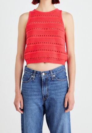 Osoba ubrana w czerwoną, bezrękawnikową, dzianą bluzkę typu crop top z wzorami ażurowymi oraz niebieskie dżinsy z wysokim stanem stojąca na jednolitym tle.