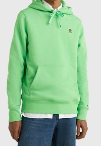 Felpa verde brillante con tasca frontale, cappuccio regolabile con cordino e logo ricamato sul petto. Materiale morbido simile al fleece.