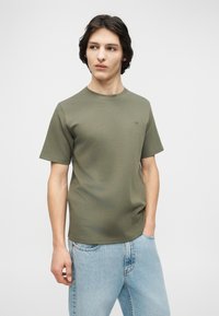 Νεαρός με σκούρα μαλλιά, φορούσε μια ελιά πράσινη κοντομάνικη μπλούζα και ανοιχτό μπλε τζιν, στέκεται και κοιτάζει προς τα αριστερά.