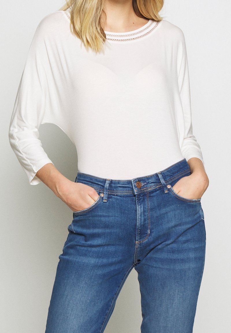 Frau in einem weißen Langarmoberteil mit Spitzenhalsauschnitt und blauen hochtaillierten Jeans, mit den Händen in den Taschen vor einem schlichten Hintergrund.