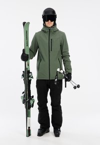 Skiudstyr i olivengrøn jakke med sorte bukser; holder grønne ski med bindinger og grå skistænger med sorte accenter. Sorte hjelme og goggles.