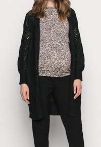 Svart stickad cardigan med öppen framsida, strukturerat mönster och långa ärmar, lagd över en rosa och svart mönstrad topp samt svarta byxor.