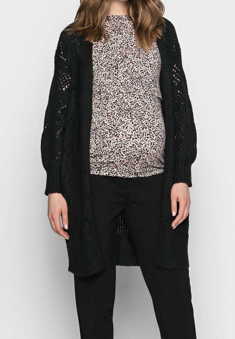 Svart stickad cardigan med öppen framsida, strukturerat mönster och långa ärmar, lagd över en rosa och svart mönstrad topp samt svarta byxor.