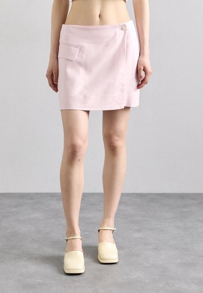 Jupe portefeuille rose avec poche et détail de bouton, texture lisse, longueur mi-cuisse, associée à des chaussures à talons jaunes claires avec brides au niveau de la cheville.