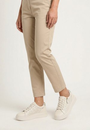 Persoon die beige slanke broek en witte veterschoenen met zijrits draagt, staand op een witte achtergrond.