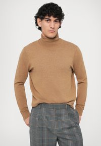 Pull à col roulé beige clair en tricot doux avec des manches longues, associé à un pantalon à carreaux gris avec des accents orange et des plis.