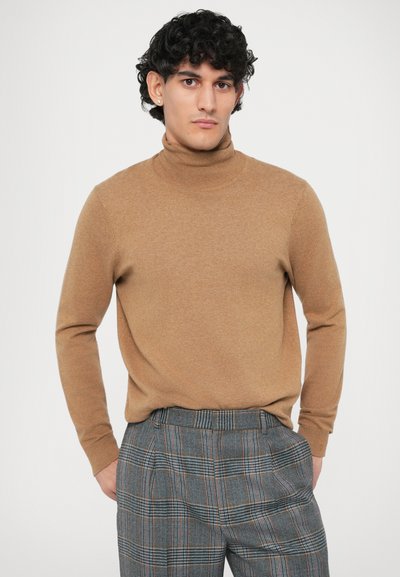 Hellbrauner Rollkragenpullover aus weichem Strick mit langen Ärmeln, kombiniert mit grauen Karohosen mit orangefarbenen Akzenten und Falten.