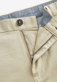 Pantaloni din bumbac bej cu fermoar și un nasture închis la culoare. Interiorul prezintă un brâu cu dungi albastre și albe. Textură netedă.
