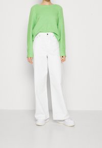 Personne portant un pull ample vert vif, un pantalon blanc à jambes larges et taille haute, et des baskets blanches, debout devant un fond uni.