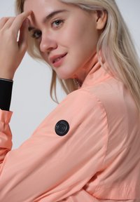 Persikka urheilu takki, jossa on pystykaulus, ribbattu musta hihansuu ja pyöreä logo-patch vasemmassa hihassa. Su smooth, kevyt kangas.