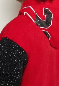 Gros plan d'une personne portant un sweat à capuche rouge avec une manche noire parsemée de taches blanches et un numéro partiel en noir et blanc audacieux sur la capuche.