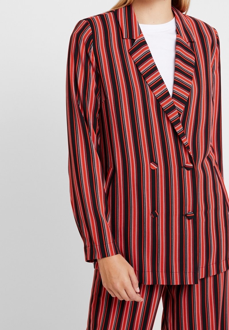 Femme portant un blazer croisé à rayures verticales rouges, noires et blanches et un pantalon assorti avec une chemise blanche en dessous.