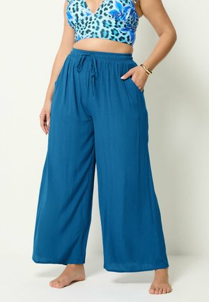 Kvinde iført løstsiddende, vidde blå bukser med snøre i taljen og blå crop top med dyreprint, stående barfodet med den ene hånd i lommen.