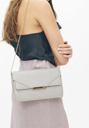 Femme portant un haut noir sans manches et une jupe claire, tenant une pochette argentée scintillante avec une bandoulière en chaîne dorée sur l'épaule.