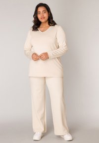 Beige, langärmeliges Oberteil mit V-Ausschnitt und passenden weit geschnittenen Hosen, mit glatter Textur. Stylisch kombiniert mit weißen Sneakers.