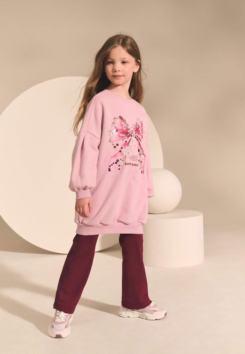 Jeune fille aux longs cheveux portant un sweat-shirt rose oversize avec un nœud en sequins, un pantalon bordeaux et des baskets roses, debout dans un décor de studio neutre.